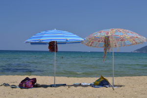 Două umbrele de plajă colorate, una albastră cu dungi și una cu modele florale, pe o plajă liniștită cu nisip fin, lângă o mare limpede, sub un cer senin.